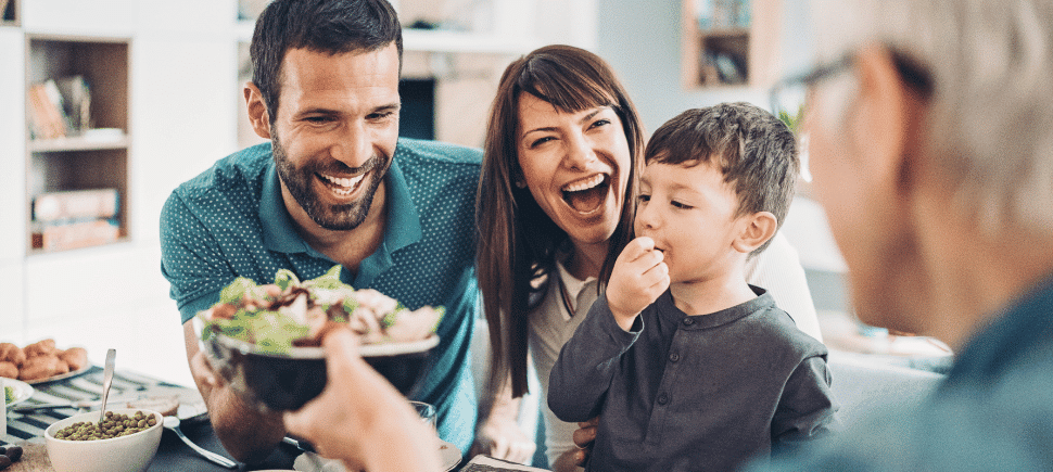 Photo of a smiling family bonding at dinner while passing a bowl of healthy salad.