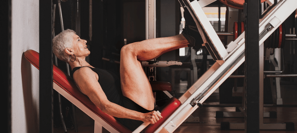 Photo of an older woman with short grey hair working out on a leg press machine at a gym