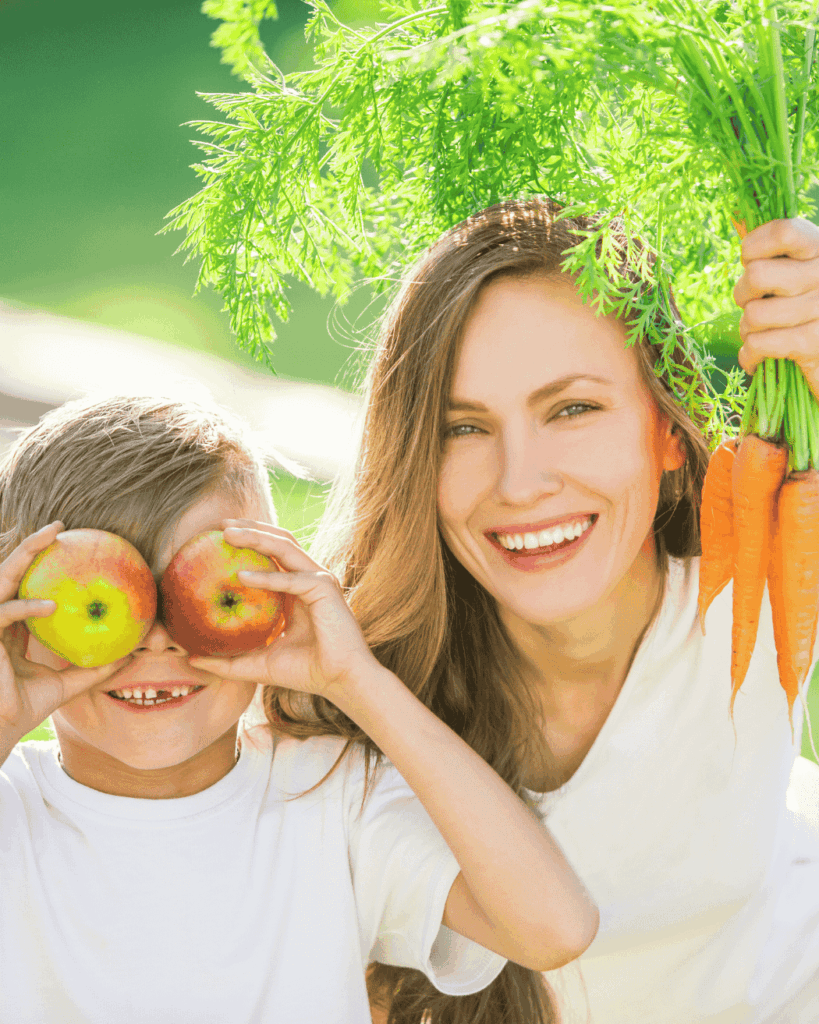 Photo of a smiling mom next to her young son. He is holding two apples in front of his eyes and she holds a bunch of carrots by the leafy tops