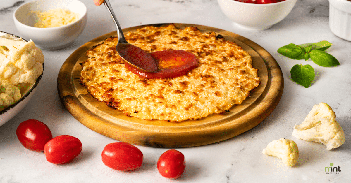 Photo of preparing a cauliflower pizza: spooning marinara sauce onto the cauliflower crust resting on a pizza stone, surrounded by fresh tomatoes, fresh cauliflower, a bowl of cheese, small bowl of marinara sauce, and fresh basil.